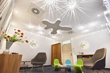 Weiß gestrichener Raum mit strahlenförmigen Lichteffekten an der Decke. Möbliert mit Holzmöbeln, bräunlichen Sesseln und Kindersesseln aus Kunststoff in Grün und Blau. 