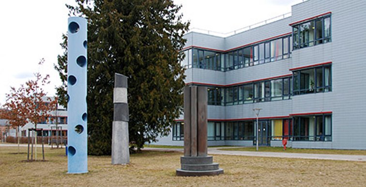 3 Säulen stehen im Dreieck zueinander, wobei sie in der Größe von links nach rechts abnehmen. Die Linke Säule ist weiß mit blauen durchgehenden  Löcher. Die mittlere besteht aus einem langen hellgrauen Abschnitt auf den versetzt drei kürzere Abschnitte in dunkel-, hell- und wieder dunkelgrau gestapelt sind. Die letzte Säule steht auf einem Sockel aus 3 Metallscheiben und besteht aus einer Hand voll Metallstangen unterschiedlicher Dicke.