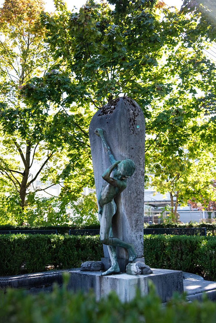 Grün angelaufene Figur eines nackten Jungen der seinen rechten Arm nach oben streckt und ein Rohr zuhält, während sein linker Arm an die eigene Schulter fasst.