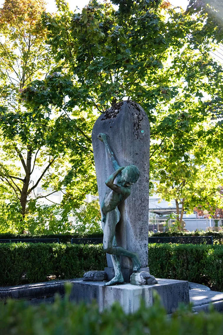 Grün angelaufene Figur eines nackten Jungen der seinen rechten Arm nach oben streckt und ein Rohr zuhält, während sein linker Arm an die eigene Schulter fasst.