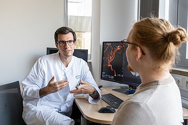 Arzt in weißer Arbeitskleidung sitzt am Schreibtisch und kommuniziert gestenreich mit einem stehenden Mann. Der Monitor auf dem Schreibtisch zeigt die radiologische Darstellung eines Blutgefäßes.