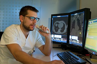 Ein Radiologe befundet Untersuchungsbilder, die auf zwei Monitoren neben ihm zu sehen sind. Er spricht in ein Mikrofon. Auf einem dritten Monitor ist die direkte Umsetzung seiner Worte in eine Textverarbeitung zu sehen.