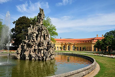 Links angeschnitten ein ovales Brunnenbecken, mit Wasserstrahl und einen kegelförmigen Felsenberg, auf dem sich verschiedene Skulpturen befinden. Im Hintergrund eine sandfarbene, halbkreisförmige, eingeschossige Orangerie mit rotem Dach.